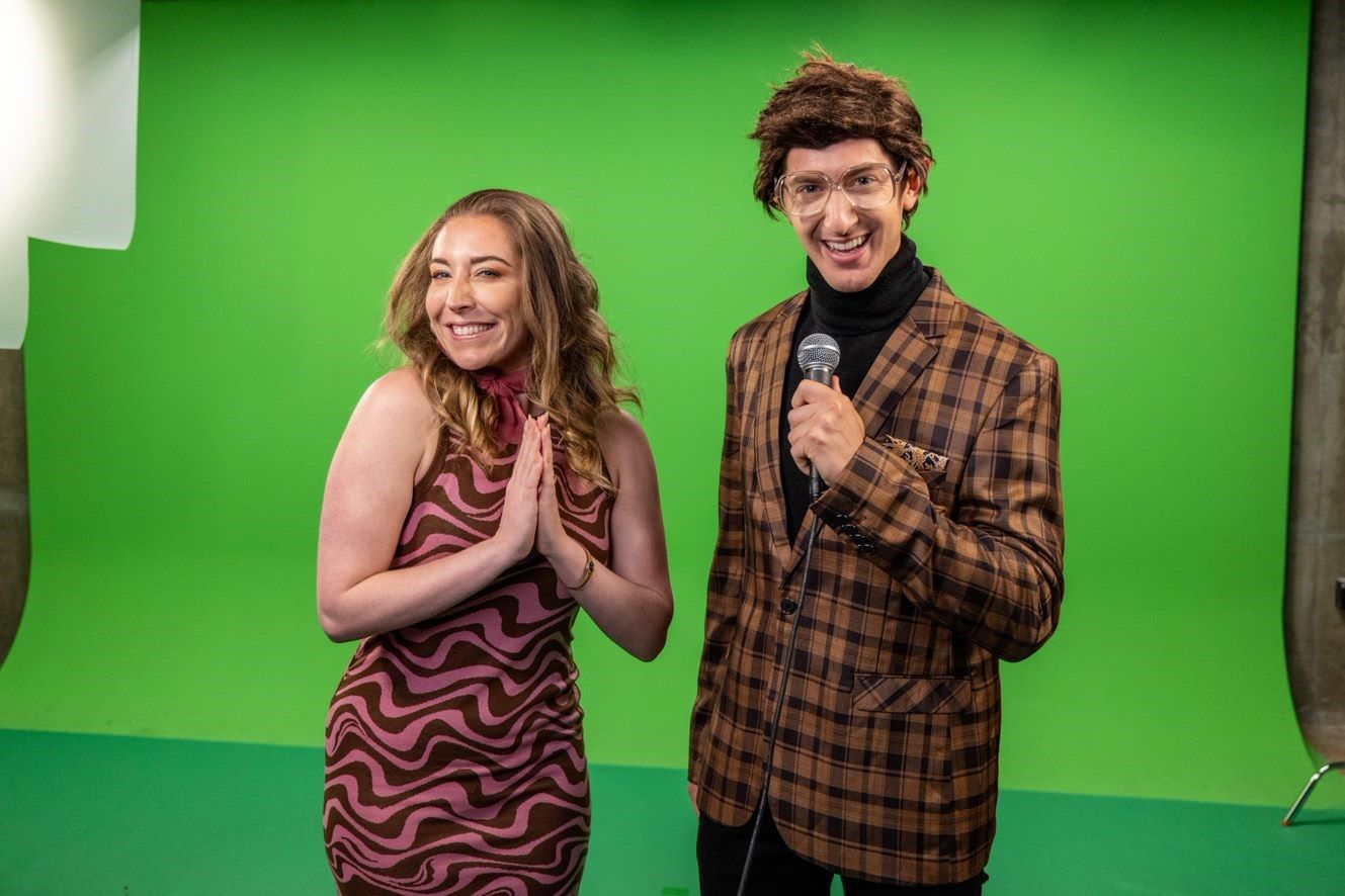 Two people pose in front of a green screen, one holding a microphone, in a lively setting.