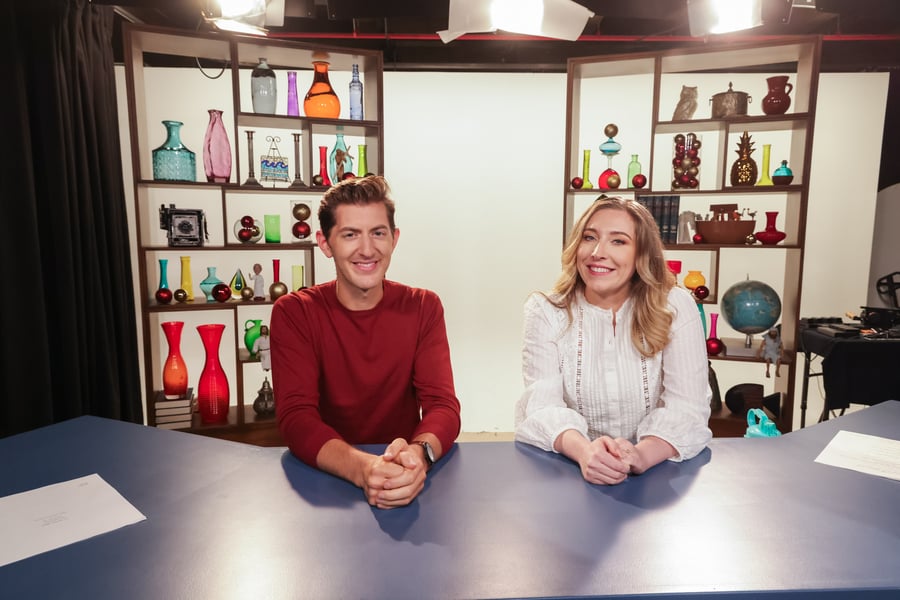 Two people are sitting at a blue desk in a studio with colorful decorations on shelves behind them.