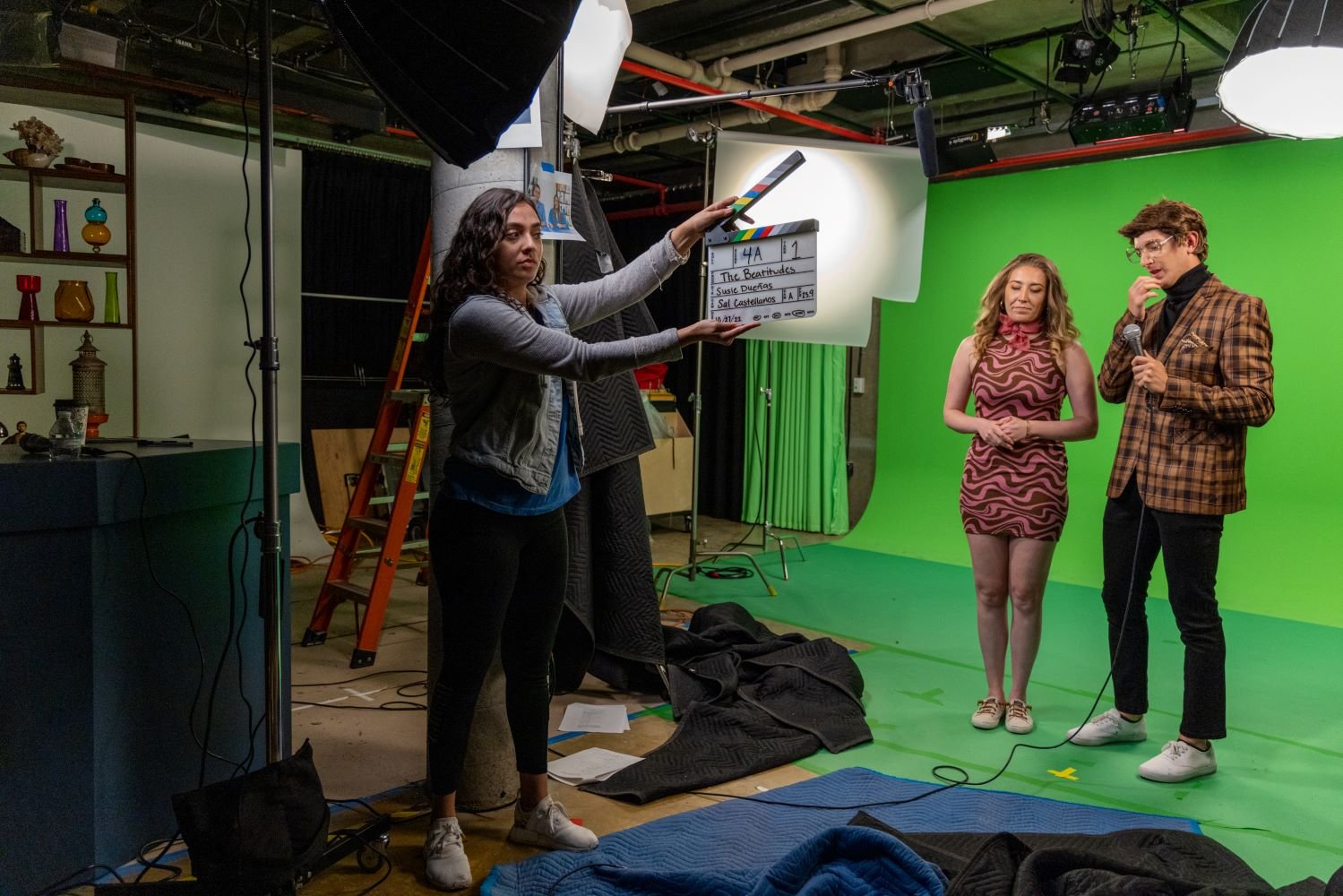 A film crew in a studio with a green screen. A person holds a clapperboard, while two others stand in front of the screen.