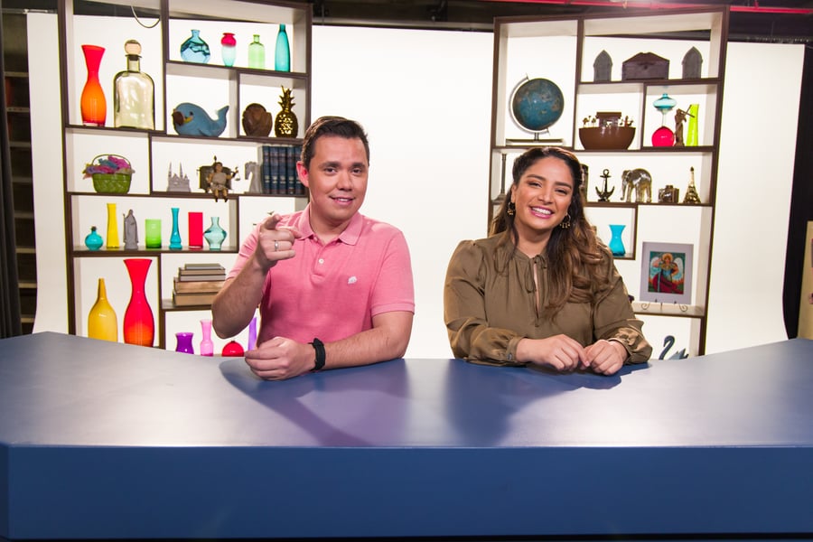 Two people are seated at a desk with colorful vases and various decorations on shelves behind them.
