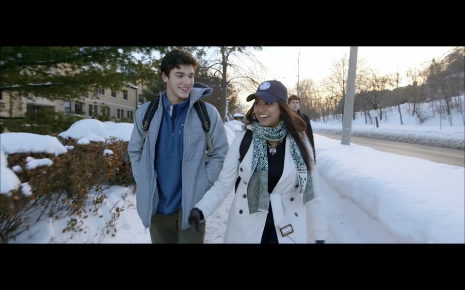 A pair of people are walking and laughing in a snowy setting, dressed warmly, with backpacks.