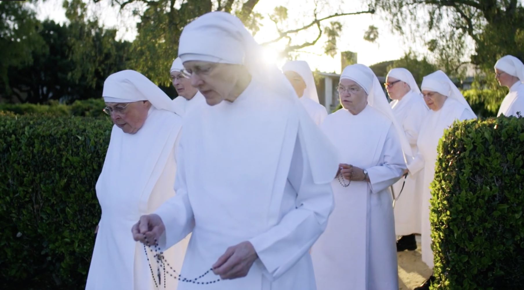 A group of nuns walks together outdoors, holding rosary beads, with the sun shining through trees in the background.