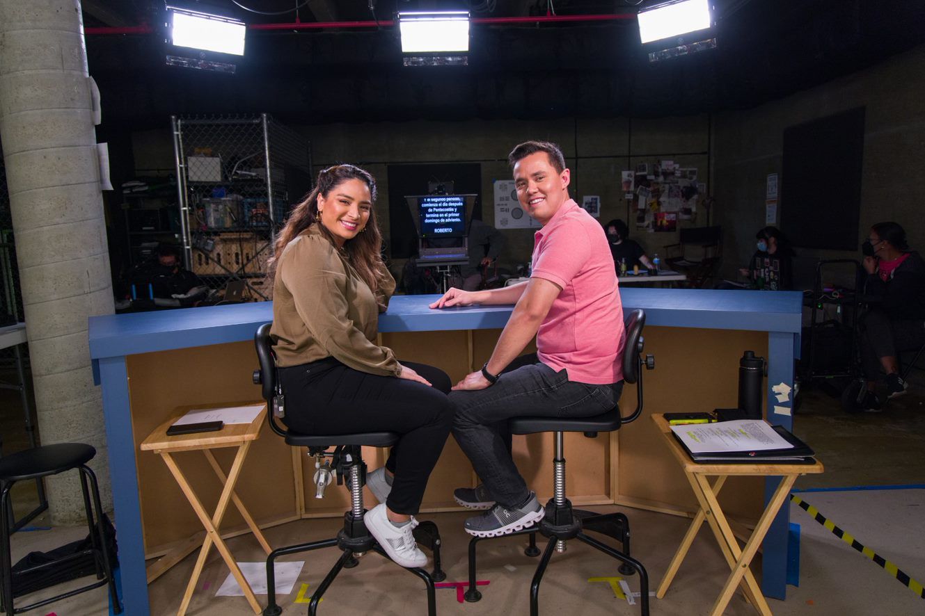 Two people are sitting in a studio with a teleprompter and lights. This relates to catholic digital media.