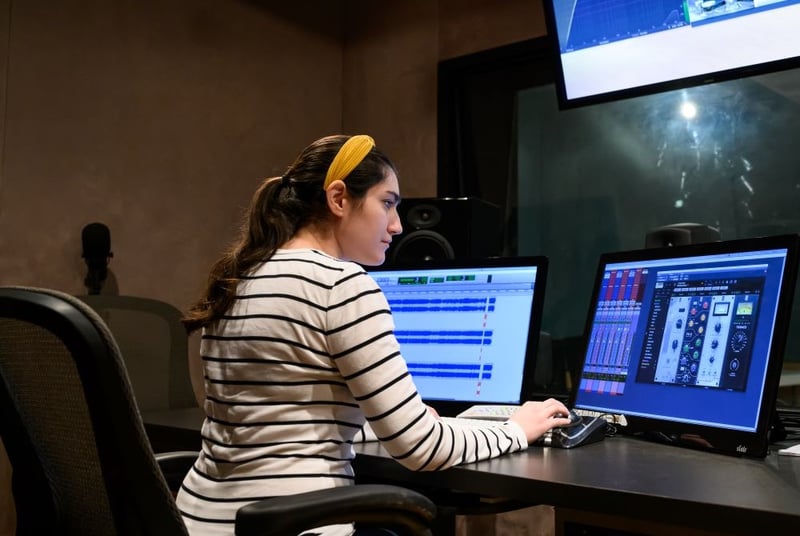 A person is working at an audio mixing station with dual monitors, possibly an audio engineer.