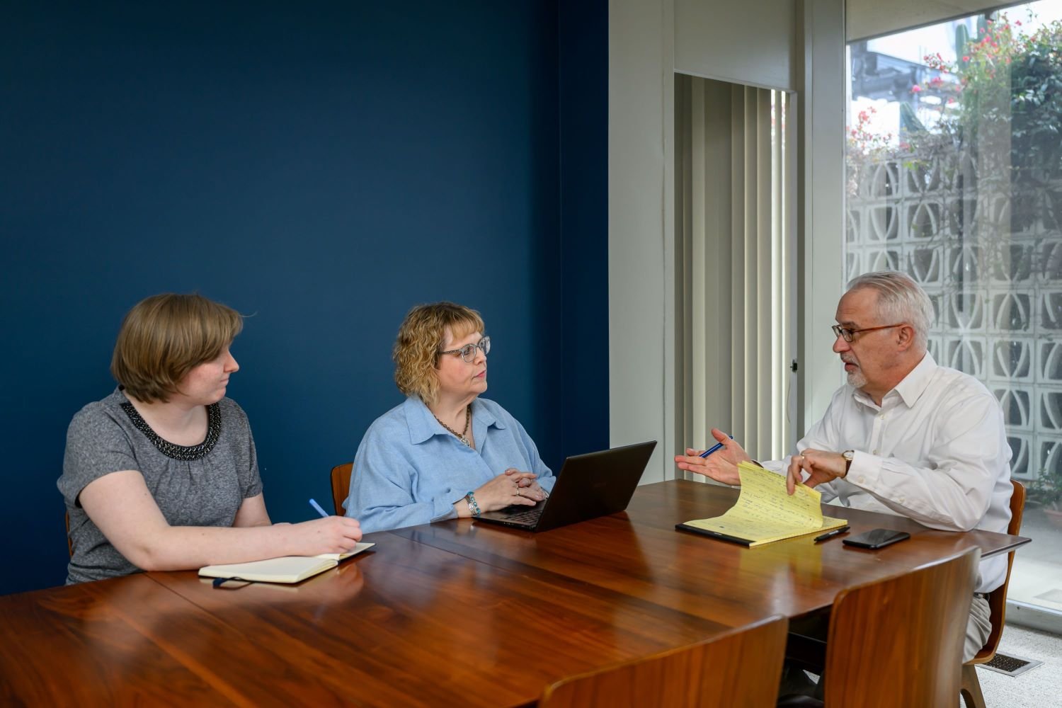 Three people are having a meeting at a wooden table with notebooks and a laptop.