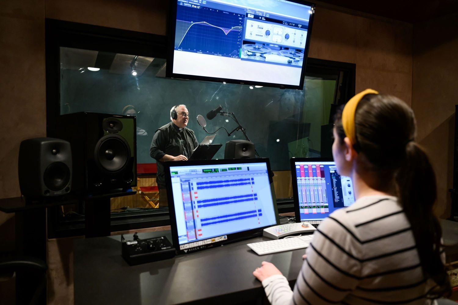 A recording studio setup with a person speaking into a microphone and another monitoring sound levels at a computer.