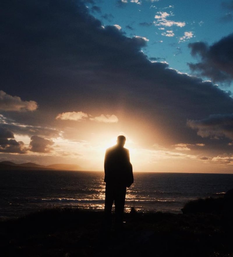A silhouette of a person standing by the sea during sunset with dramatic clouds in the sky.
